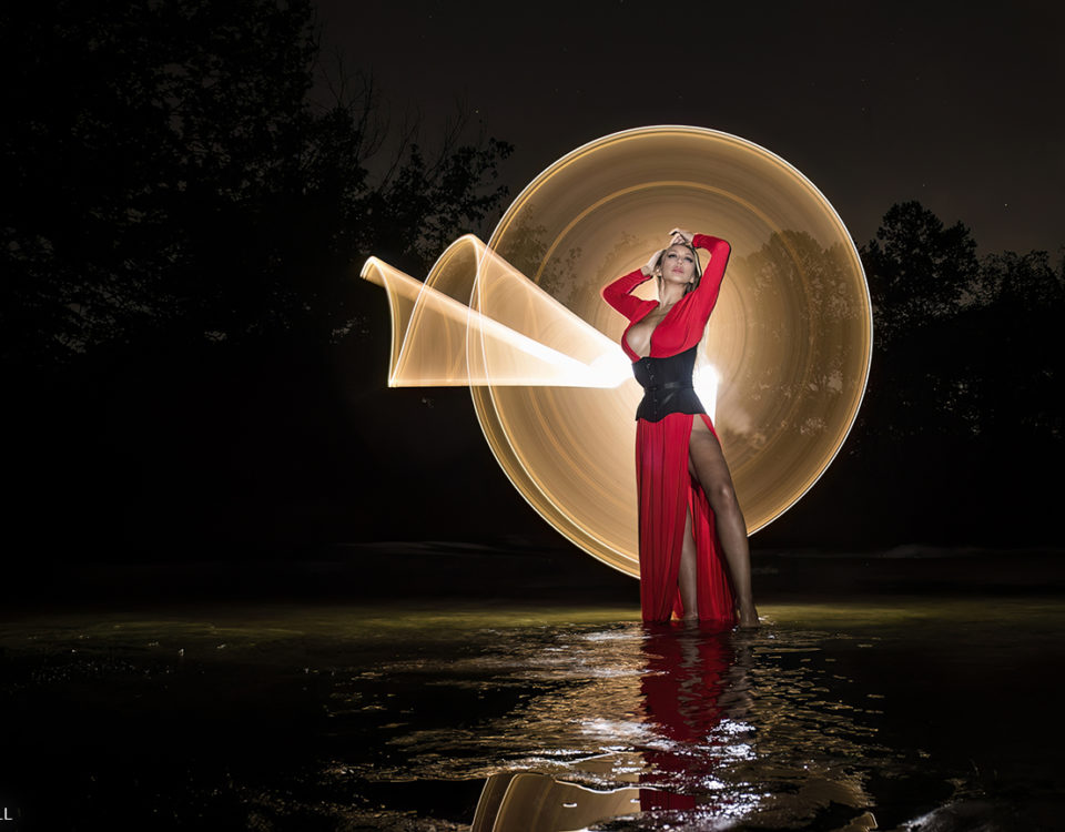 Model, Harlow Wilde, in red dress and black corset, standing in a creek in Smyrna Tennessee, a light painting circle behind her.